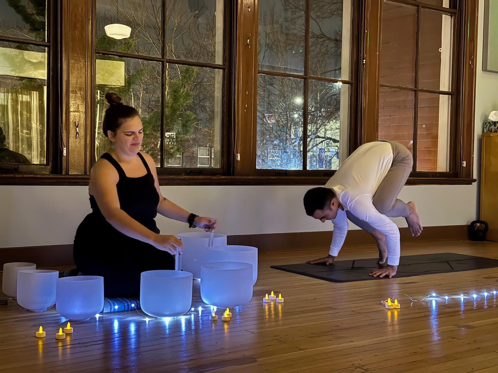 In a candlelit studio with tall wood-framed windows, a practitioner plays crystal singing bowls while a man holds crow pose on a yoga mat
